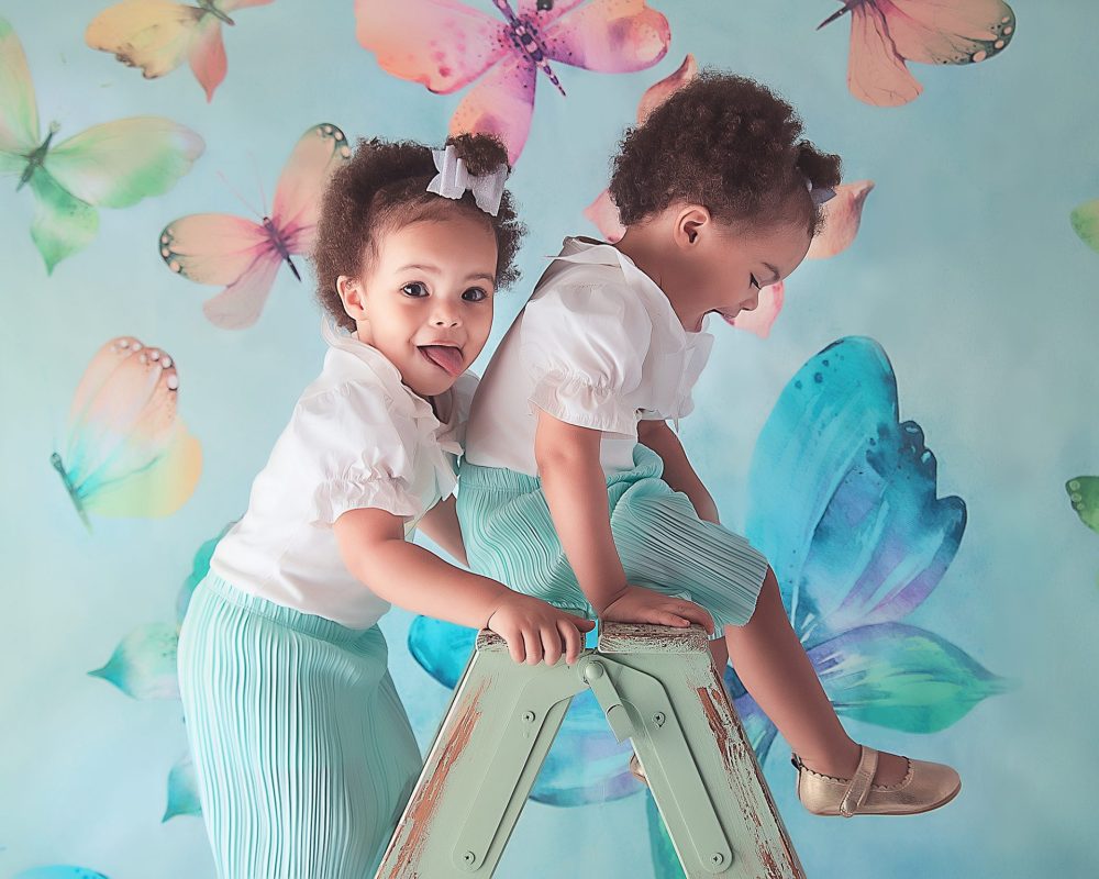Spring Mini Session, Discounted Photoshoot at the Baby Boutique Manchester. Bring your little one's for a fun spring photoshoot at a discounted price. Two little girls climbing a green ladder, wearing matching outfits. Against a butterfly backdrop in green and blue tones.