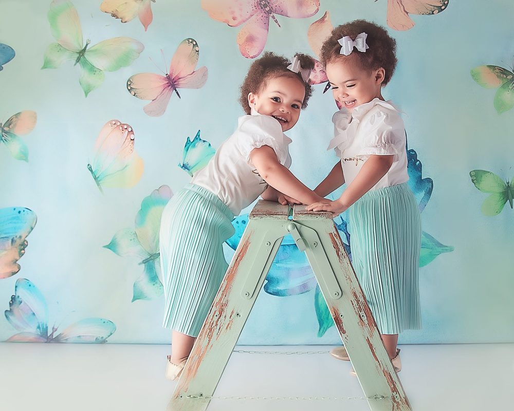 Spring Mini Session, Discounted Photoshoot at the Baby Boutique Manchester. Bring your little one's for a fun spring photoshoot at a discounted price. Two little girls climbing a green ladder, wearing matching outfits. Against a butterfly backdrop in greens and blue tones.