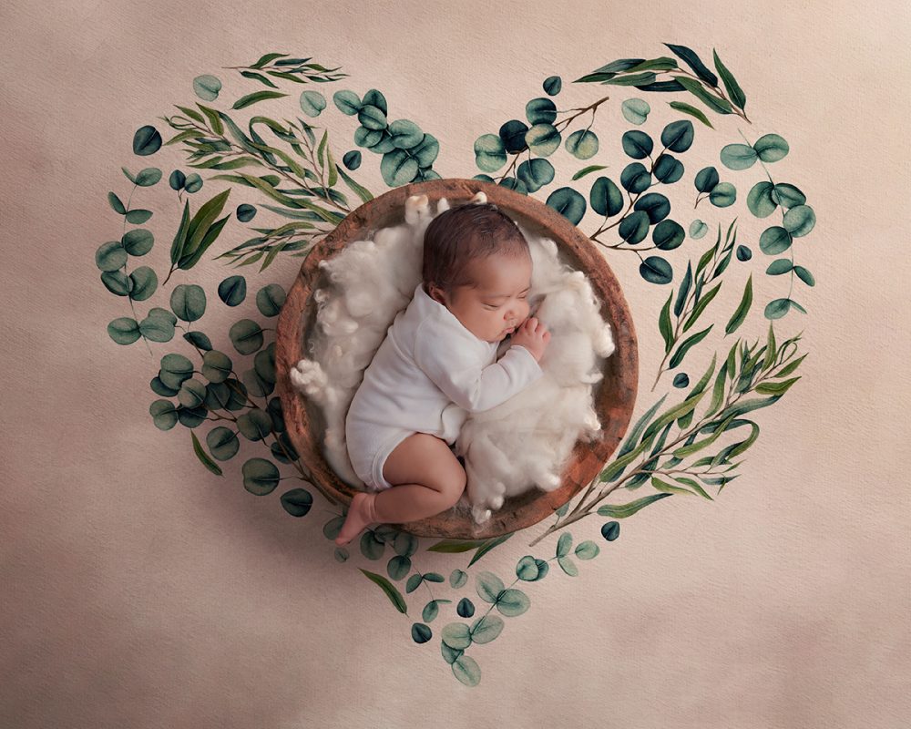 Newborn baby in a composite image taken by Baby Boutique Manchester. Newborn photography studio. Baby lies in a brown bowl asleep surrounded by green watercolour leaves.