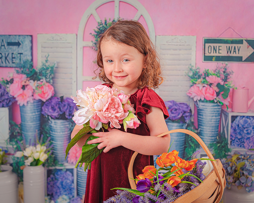 Spring Mini Session, Discounted Photoshoot at the Baby Boutique Manchester. Bring your little one's for a fun spring photoshoot at a discounted price. Little girl standing against a bright coloured floral backdrop. Holding a basket with flowers and a bouquet of pink flowers. Looking and smiling at the camera.