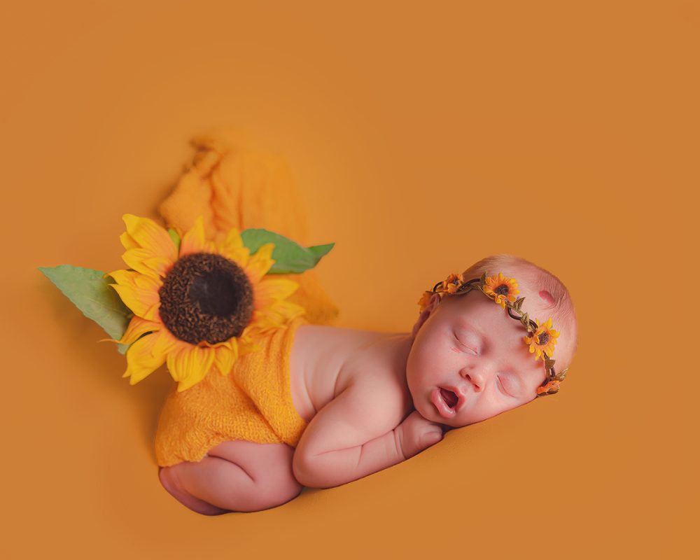 Little baby lying on a yellow backdrop fast asleep. She has a sunflower crown on and is lying on her front.