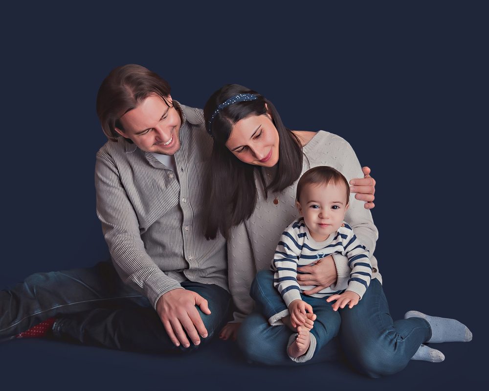 The image was taken at the Baby Boutique Manchester. Featuring a Dad and Mum with their little baby.  Positioned against a dark blue backdrop. Wearing neutral colours and blues. The parents are looking down at their baby.