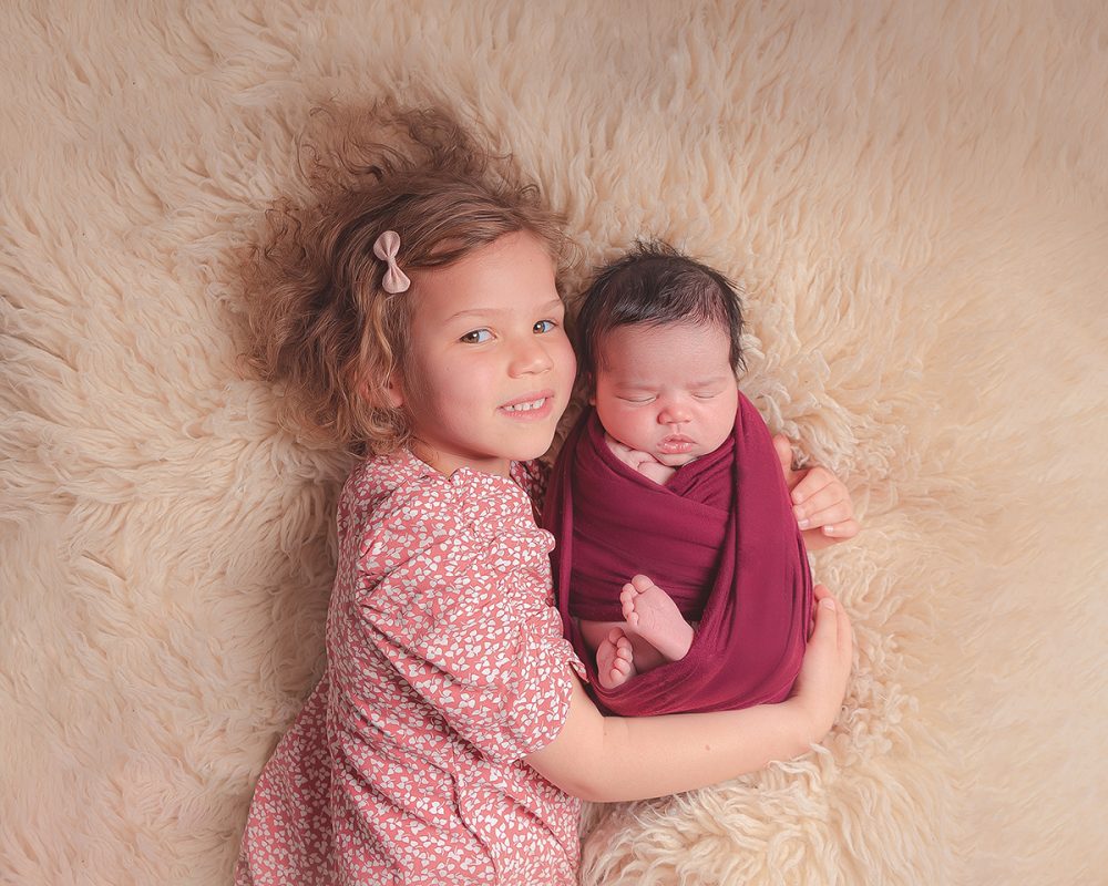 Two little girls lying on a fluffy blanket. The oldest child is looking up at the camera and has a bow in her hair. The newborn baby is wrapped tightly in a red swaddle. She is a sleep with her hands near her faced. Image taken at the Baby Boutique Manchester.