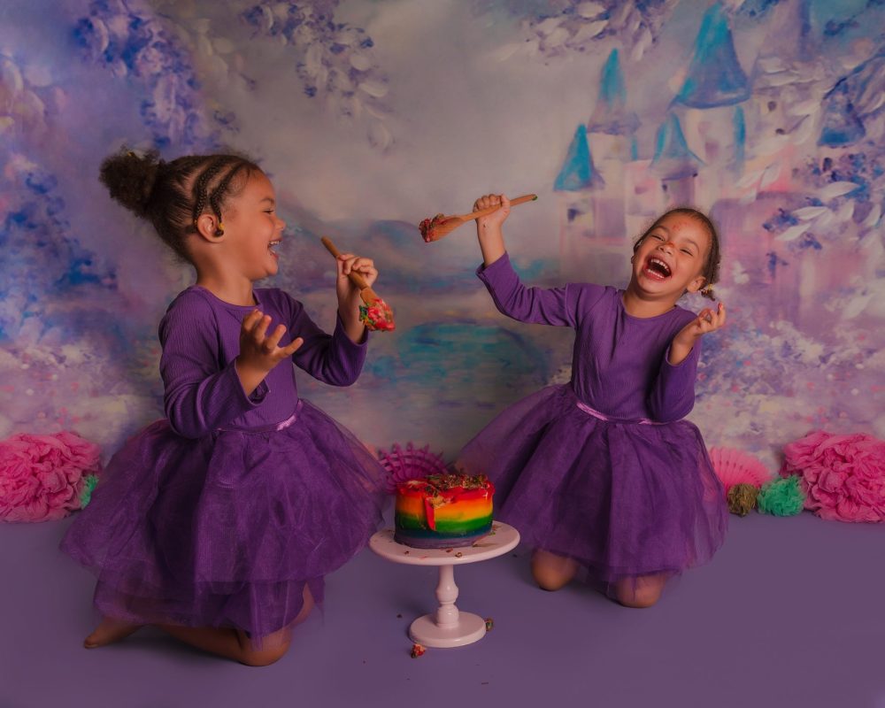 Colourful photoshoot with twins and a rainbow cake smash. They are having a cake food fight and laughing. Both wearing purple.