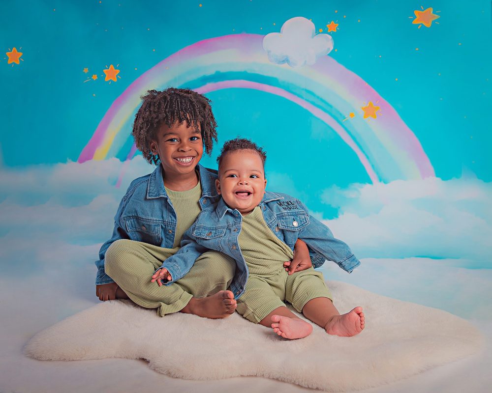 Two little boys sitting on a cream fluffy rug against the blue backdrop that features a rainbow and stars. Wearing matching green outfits and denim jackets.