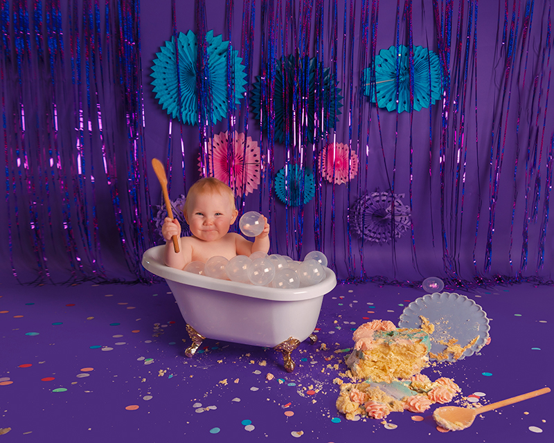 Photograph showing our new Bubble Bath Prop against a dark purple backdrop. There is a little girl sitting in a white bubble bath with gold legs. Holding a spoon and playing with some plastic clear balls. There is a cake smashed on the floor. 
