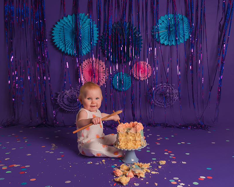 Photograph showing a cake smash against a dark purple backdrop. There is a little girl wearing a white outfit scattered with pink flowers. She is hitting a wooden spoon against a cake. The cake is blue and purple with pink piping. Taken at the Baby Boutique Photography Studio.
